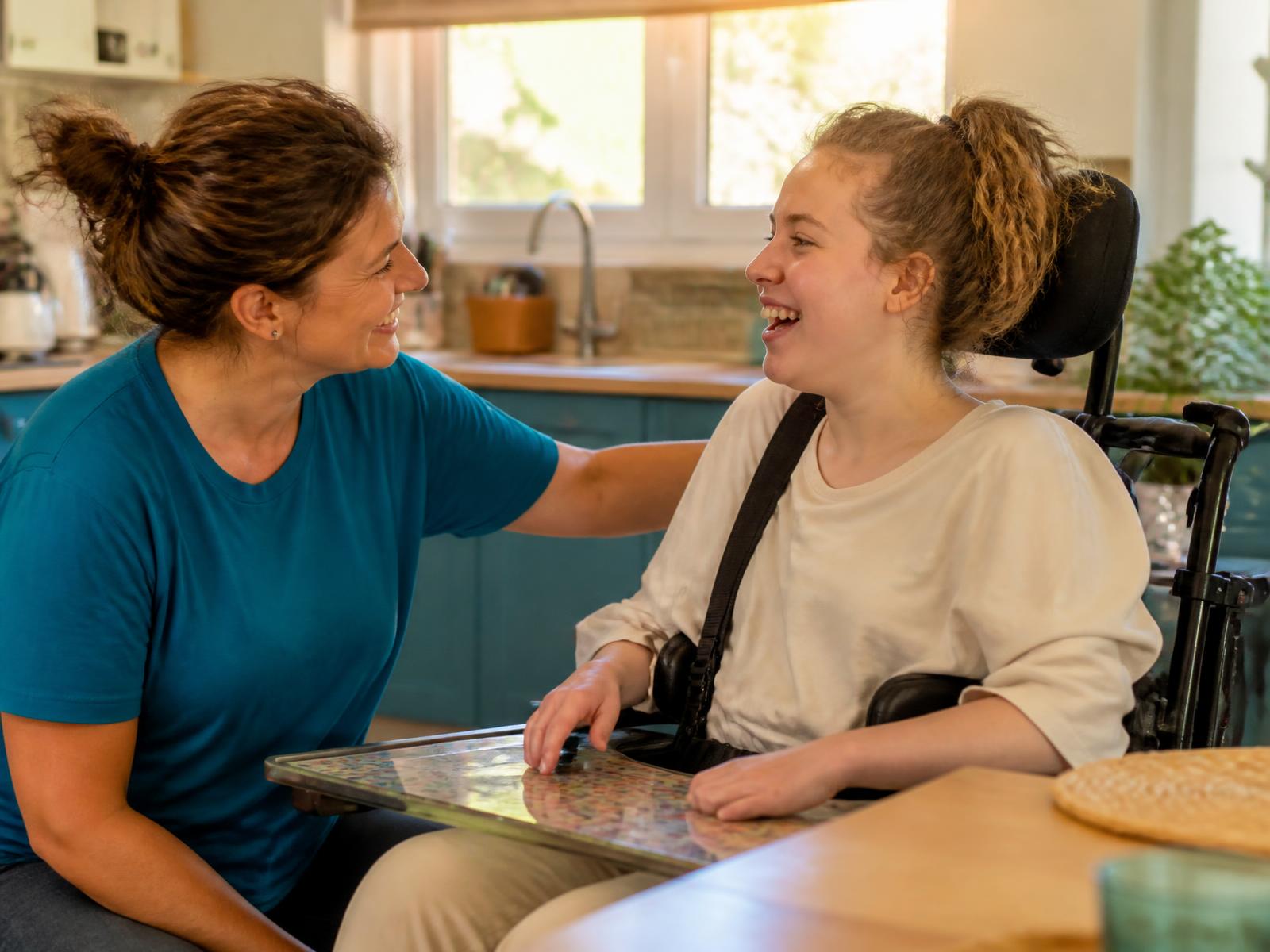 A support worker and a young woman who uses a wheelchair laughing together in a sunlit home kitchen.